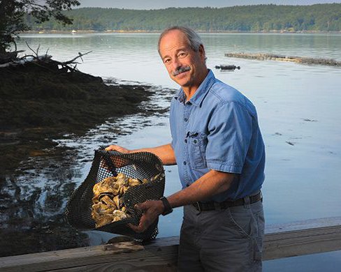  A man stands next to a body of water while holding a net full of shellfish. 