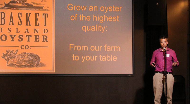  A man stands in front of a microphone on stage. A large projection screen behind him shows business messaging. 