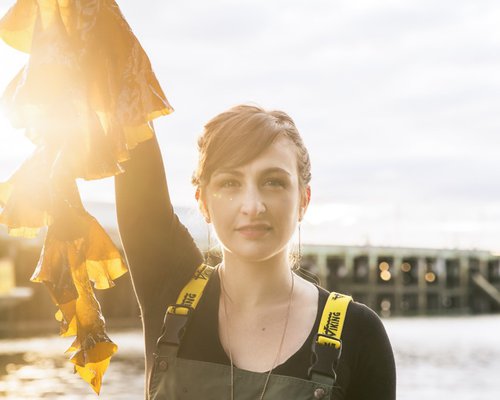  A young woman wearing work overalls holds a piece of seaweed overhead. 